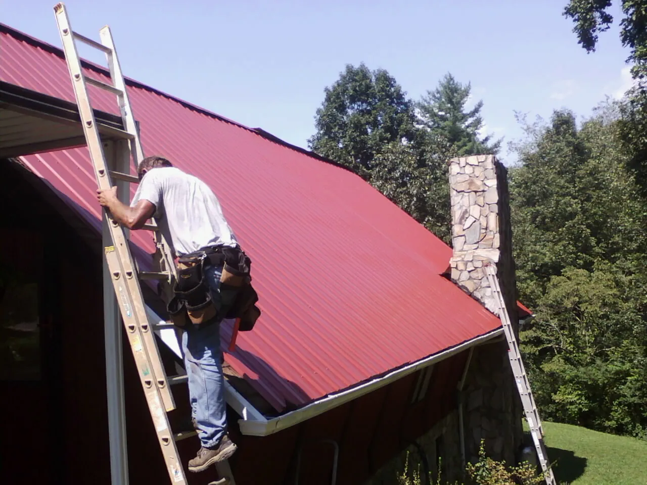 Roofer on a ladder beside a red metal roof and stone chimney — Soffit & Fascia Repair by Roofing Brothers in Bannock